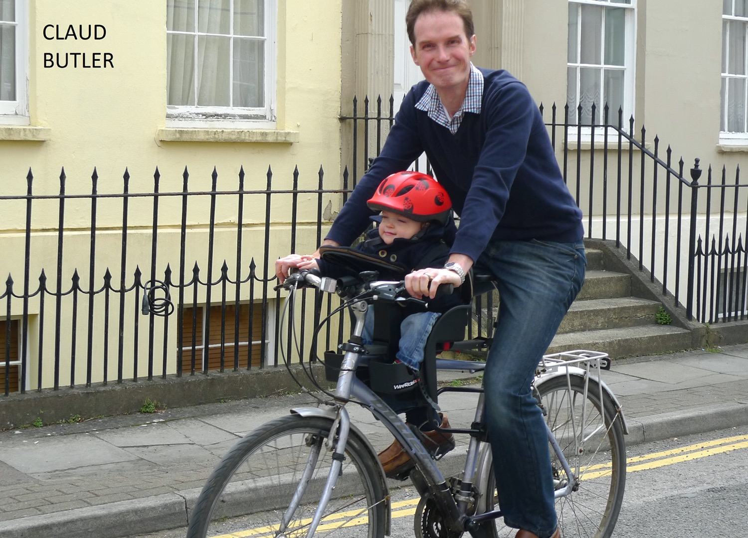 Father cycling on residential street with toddler in centre-mounted WeeRide bike seat, child wearing red helmet and grinning