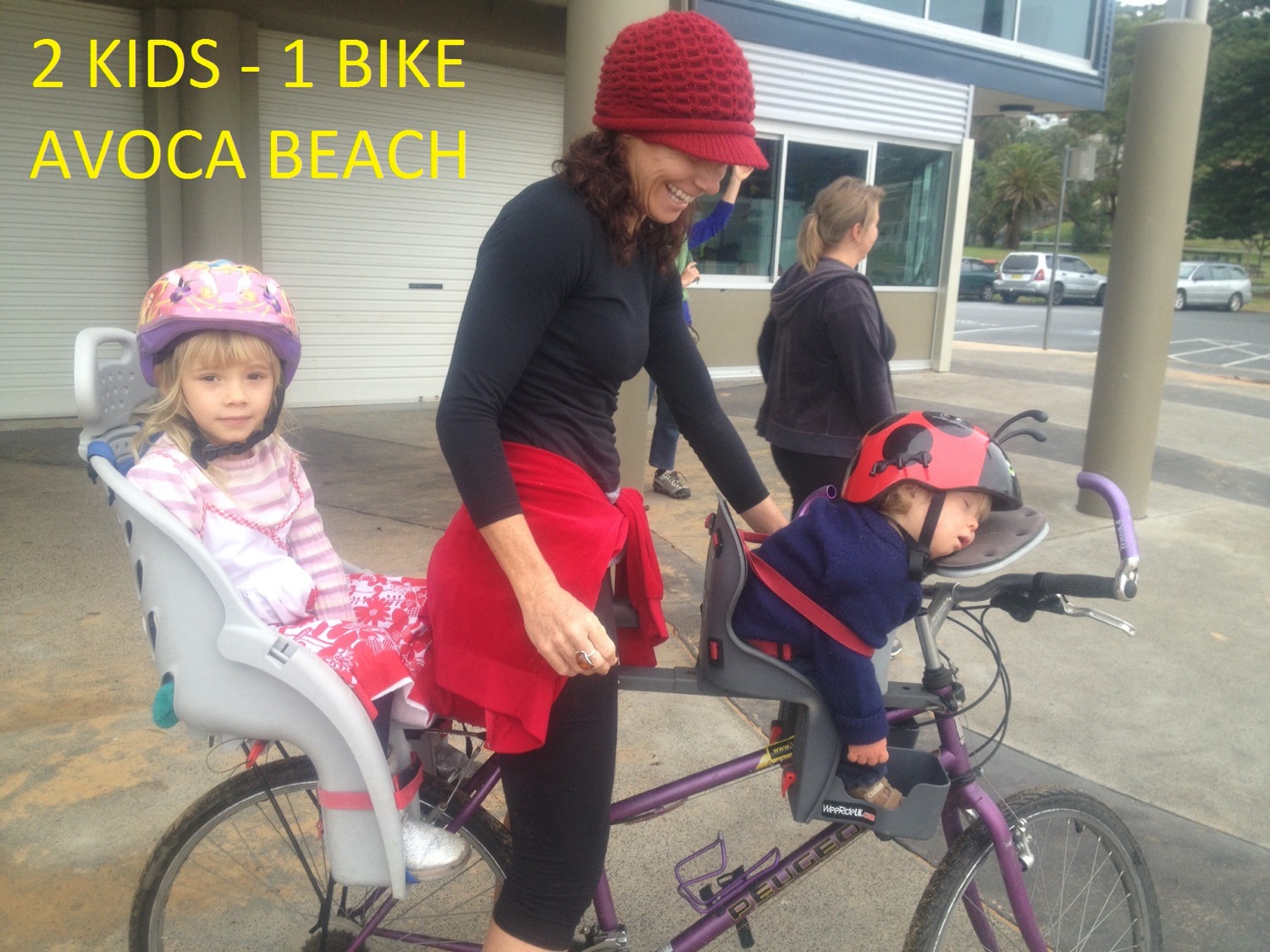 Mother at beach with two children on one bike — toddler asleep in front-mounted WeeRide seat, older child in rear seat
