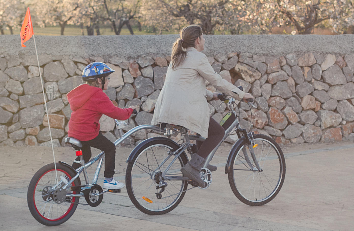 Mother and child cycling together on a scenic stone-walled path, child riding a WeeRide Co Pilot tag-along with helmet and safety flag