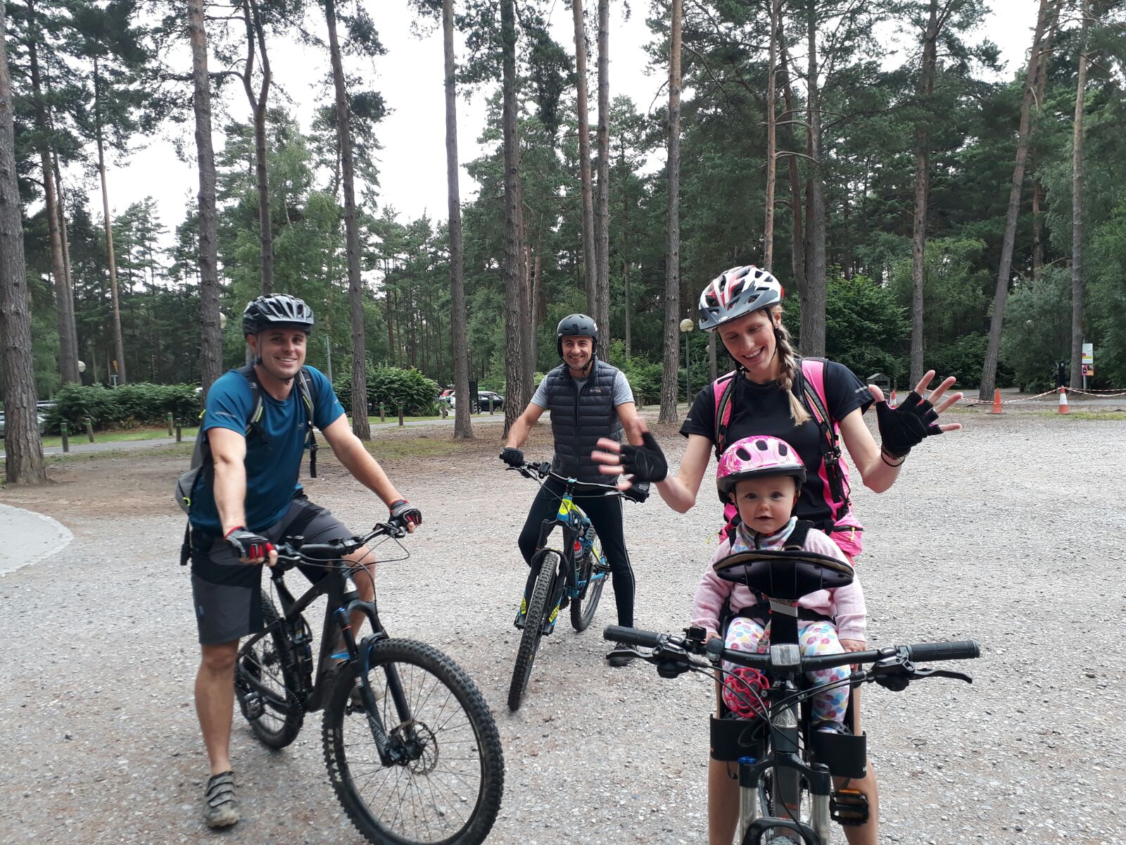Group of adults and baby pausing on a forest trail — baby in front-mounted WeeRide seat wearing pink helmet, mum celebrating with hands raised